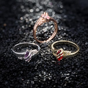 Op de afbeelding: Drie ringen met edelsteenaccenten worden getoond op een donker, getextureerd oppervlak. De ringen zijn goud, zilver en roségoud. Elke ring heeft een verschillend gekleurde edelsteen en het jaar "2025" is gegraveerd.