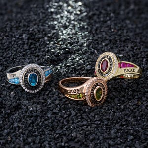 May include: Three class rings with oval center stones and surrounding text. One ring is silver with a blue stone and the year 2026. Another is gold with a red stone and the name "Brad". The third is rose gold with a green stone and the year 2025.