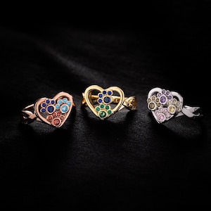 May include: Three heart-shaped rings featuring paw print designs. The left ring is rose gold with blue and red stones. The center ring is gold with blue and green stones. The right ring is silver with purple, pink, and yellow stones.