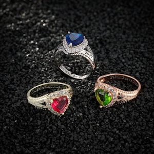 May include: Three rings with heart-shaped gemstones in silver, gold, and rose gold. The rings feature red, blue, and green gemstones, surrounded by clear stones. Each ring has engraved text, with one reading "2027".