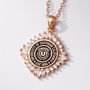 May include: Rose gold necklace with a pendant featuring the University of Southern California seal. The pendant is round with a black and gold design, surrounded by clear crystals. The chain is delicate and matches the pendant's color.