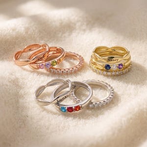 Pode incluir: Três conjuntos de anéis empilháveis em tons de ouro rosa, ouro amarelo e prata. Cada conjunto inclui uma banda lisa, uma banda gravada com nomes como Dad, Nancy ou Luna, e uma banda com três pequenas pedras preciosas coloridas e claras.