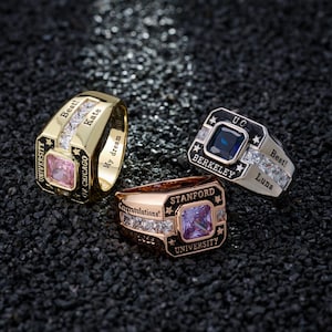 May include: Three university class rings with square gemstones. One is gold with a pink stone, another is silver with a blue stone, and the third is rose gold with a purple stone. Each ring has text, including "Best!" and university names.