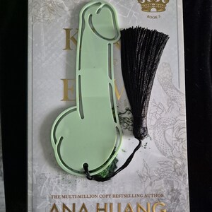May include: A pale green phallic bookmark with a black tassel, resting on a book. The book cover features the title "Kings of Sin" and the author's name, Ana Huang, in gold lettering.
