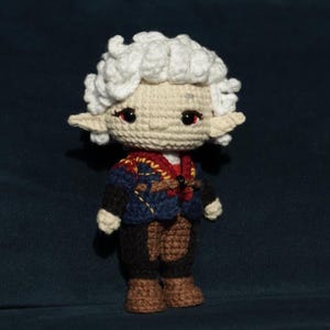 Può includere: Bambola all'uncinetto fatta a mano con viso color crema, capelli ricci bianchi e orecchie a punta. La bambola indossa una giacca blu con dettagli dorati, pantaloni marroni e stivali marroni. Gli occhi sono rossi. Un oggetto unico fatto a mano.