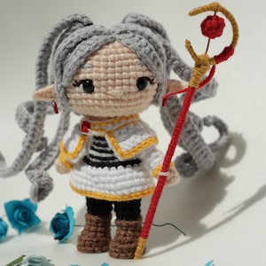 Puede incluir: Muñeca de ganchillo hecha a mano con cabello gris, vestida con un atuendo blanco y negro con ribetes amarillos. La muñeca tiene ojos grandes y sostiene un bastón rojo y dorado con una flor roja en la parte superior. Flores de papel azules esparcidas.