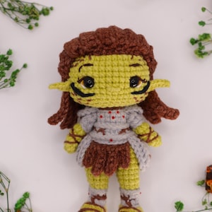 May include: A crochet doll with light green skin, pointed ears, and brown hair. The figure wears a grey chest piece with red accents, a brown skirt, and brown leg wraps. It has black eyes and black markings under the eyes on its face.