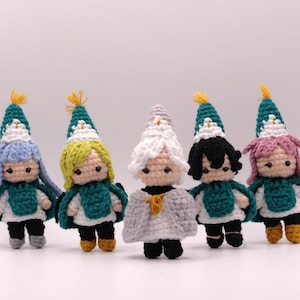 Puede incluir: Cinco pequeñas muñecas de ganchillo dispuestas en fila. Cada muñeca lleva un gorro puntiagudo con una borla amarilla y una capa verde sobre un conjunto blanco. Las muñecas tienen diferentes colores de pelo: azul claro, amarillo, blanco, negro y rosa, con ojos negros.