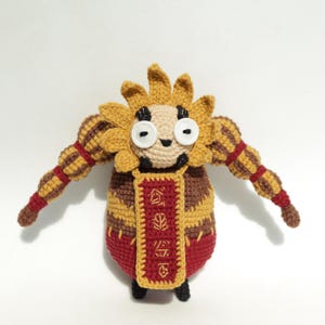 May include: A handmade crochet doll with a whimsical design. The doll features a yellow sunflower-like head, button eyes, and striped arms. The body is a combination of red, brown, and yellow, with a decorative banner. The doll has black feet.