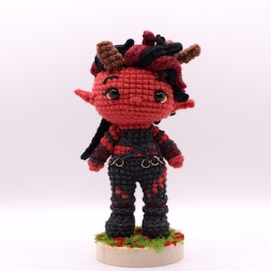 May include: A handmade crochet doll with a red face, black hair, and brown horns. The doll wears a black and red outfit with gold-colored accents. It stands on a wooden base with green moss and red accents.