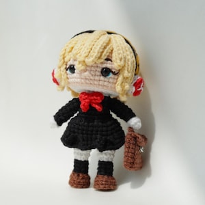 May include: Crocheted doll with blonde hair, wearing a black dress, red bow, and brown shoes. The doll has a small brown bag and is handmade. The doll's eyes are blue and it has a white collar and cuffs.