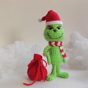 May include: A crocheted green character wearing a red and white striped scarf and a red Santa hat with a white pom-pom. A red drawstring bag sits nearby. The figure has a smiling expression and is a Christmas-themed item.