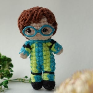 May include: A handmade crochet doll with brown hair and blue glasses. The doll wears a blue and green striped outfit with black accents. The doll's black shoes complete the look. This unique doll is a charming handmade item.