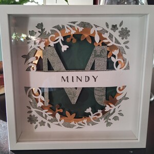 May include: A white framed shadow box with a layered design. The design features a large letter "M" in a textured silver color, with the name "MINDY" below. The design is surrounded by white and gold floral cutouts, set against a dark green background.