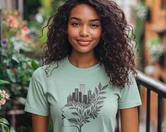 Puede incluir: Una camiseta verde claro con un diseño gráfico. El diseño presenta un paisaje urbano dentro de una silueta de árbol en forma de rostro, en tonos grises y negros. La persona de la imagen tiene cabello rizado oscuro y está sonriendo.