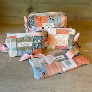 May include: Four quilted fabric pouches in various sizes, featuring a patchwork design with floral and geometric patterns in shades of peach, blue, and green. Each pouch has a zipper closure and a small label with text. The pouches are arranged on a wooden surface.