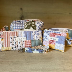 May include: A collection of patchwork fabric accessories, including a zippered pouch, a rectangular wallet, a small coin purse, and a wristlet key fob. The items feature floral, gingham, and striped patterns in various colors. Each item has a label that reads "Eva Hinkel".