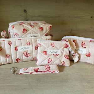May include: A collection of quilted, light pink cosmetic bags and accessories, patterned with red strawberries. The set includes a rectangular bag, a zippered pouch, a small square pouch, and a wristlet keychain. Each item has a white label with text.