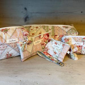 May include: A collection of floral patchwork fabric bags and pouches in various sizes. The bags feature a cream background with red and pink roses, green leaves, and a white zipper. Each bag has a small label with text.