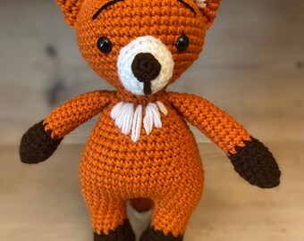 May include: A handmade orange crochet fox stuffed animal with brown accents. The fox has a white chest, white ear details, and black eyes. The fox is standing upright with its arms and legs extended.