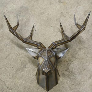 Puede incluir: Escultura geométrica de cabeza de ciervo en metal con cuernos puntiagudos. La escultura está hecha de piezas de metal soldadas, creando un diseño facetado. El color es una mezcla de plata y marrón, con marcas de soldadura visibles. Arte mural moderno.