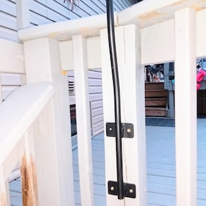 Puede incluir: Un soporte de mástil de bandera de metal negro montado en una barandilla de terraza de madera blanca. El soporte tiene dos soportes metálicos con tornillos.