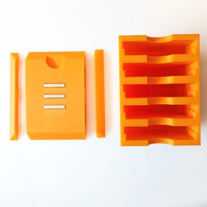 May include: Bright orange, modular storage system. The set includes a rectangular box with three silver bars, two long, thin pieces, and a multi-tiered organizer. The organizer has five shelves with curved cutouts. All pieces are made of a solid, opaque material.