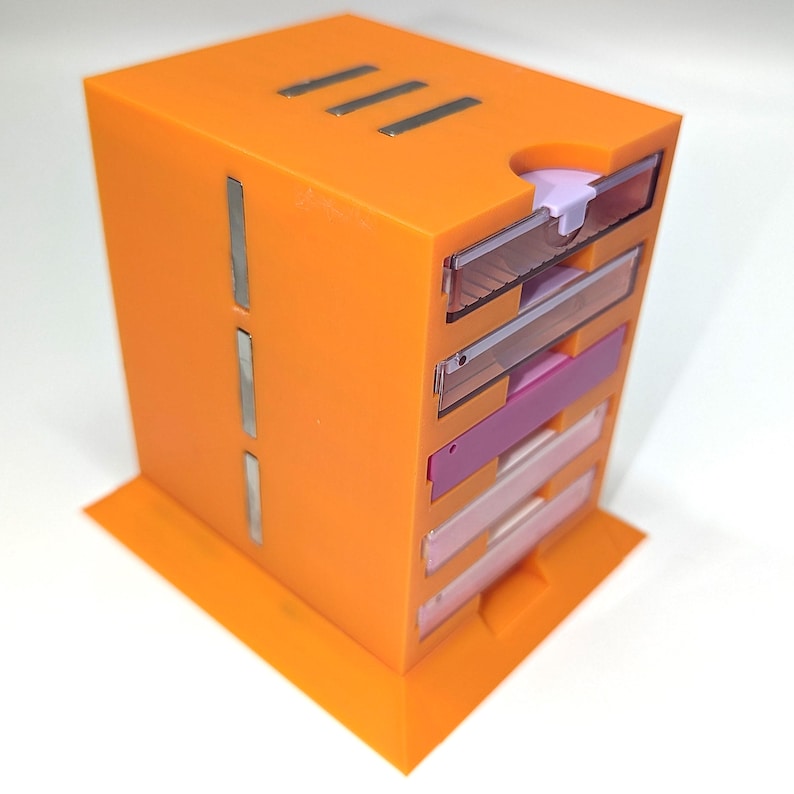 May include: An orange, multi-drawer storage unit with a rectangular shape. The unit has a series of clear and colored drawers, including pink and white. The top features three parallel silver lines and a small notch. The unit sits on an orange base.