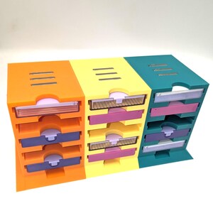 May include: A set of three colorful desk organizers. Each organizer has three pull-out drawers in orange, yellow, and teal. The drawers are accented with purple and pink. The top of each organizer has three horizontal slits.