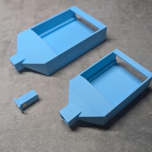 May include: Three light blue 3D-printed objects. Two are rectangular with a sloped spout and a raised rim. A smaller, rectangular piece is also present. The objects are on a gray surface.