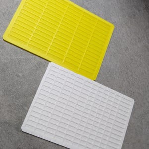 May include: Two rectangular trays. One is bright yellow, the other is white. Both have a grid-like pattern of small, raised rectangles. The trays are likely made of plastic and are designed for organizing small items or components.