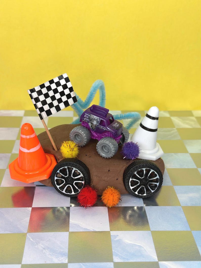 Op de afbeelding: Een kleine paarse monstertruck met grote grijze banden, een geblokte vlag en oranje en witte verkeerskegels. De truck is versierd met kleurrijke pompons en staat op een bruin oppervlak, met een gele achtergrond.