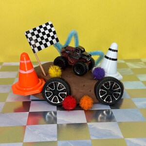 Op de afbeelding: Een miniatuur monstertruck met zwarte wielen en een rood-zwarte carrosserie staat op een bruine heuvel, naast een geblokte vlag en een oranje verkeerskegel. Extra elementen zijn twee reservewielen, pompons en een wit en zwart gestreepte kegel.