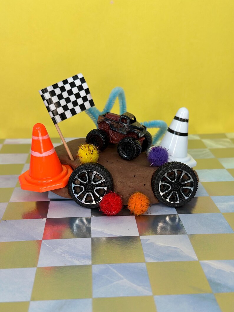 Op de afbeelding: Een miniatuur monstertruck met zwarte wielen en een rood-zwarte carrosserie staat op een bruine heuvel, naast een geblokte vlag en een oranje verkeerskegel. Extra elementen zijn twee reservewielen, pompons en een wit en zwart gestreepte kegel.