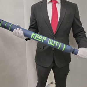 Könnte beinhalten: Ein blau-schwarzer Baseballschläger mit grünen Akzenten und dem Schriftzug "KEEP OUT)" in Grün. Der Schläger hat eine zylindrische Form mit grünen Ringen und einem strukturierten Griff. Der Schläger wird von einer Person in einem schwarzen Anzug, weißen Handschuhen und einer roten Krawatte gehalten.