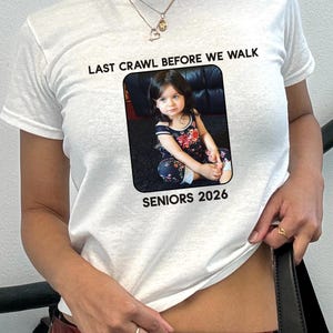 May include: White t-shirt with the text "LAST CRAWL BEFORE WE WALK" above a framed photo of a child. The photo is labeled "SENIORS 2026". The shirt is short-sleeved.