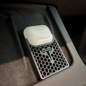 May include: A black and gray 3D-printed holder for wireless earbuds, featuring a honeycomb pattern and the Tesla logo. The holder is designed to fit in a car's center console, with a white earbud case resting inside.