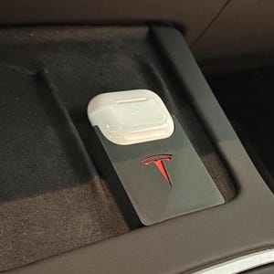 Könnte beinhalten: Ein weißes AirPods-Etui liegt auf einem schwarzen Halter mit rotem Tesla-Logo. Der Halter befindet sich in der Mittelkonsole eines Autos mit dunklem Innenraum.