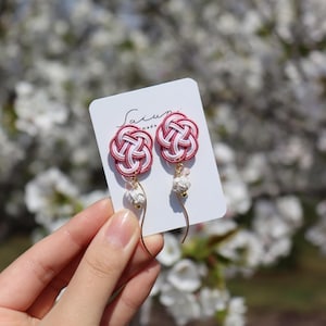 Puede incluir: Un par de pendientes rosa y blancos con un delicado diseño de nudo y detalles de perlas. Los pendientes se muestran en una tarjeta blanca con el texto "Saiun Made".
