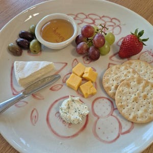 May include: A white ceramic plate with a selection of appetizers. The plate features olives, grapes, cheese, crackers, honey, and a strawberry. A small knife is next to a wedge of brie cheese. The plate has a decorative red design.