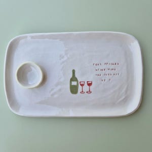 May include: A rectangular off-white ceramic serving tray with rounded corners and an attached small circular bowl. The tray features a green wine bottle illustration, two red wine glasses, and the text "real friends bring wine and fuck off by 9".