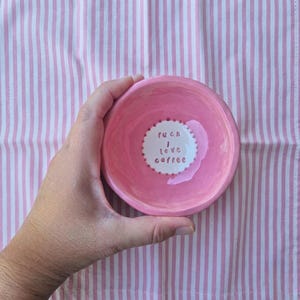 May include: A small, round, pink ceramic dish with the text "fuck I love coffee" in a white circle. The dish is held in a hand against a pink and white striped background.
