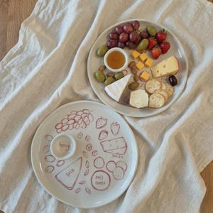 May include: Two white ceramic serving platters, one with grapes, strawberries, cheese, crackers, olives, and honey. The other platter has a hand-drawn design of food items, including grapes, strawberries, and cheese, with the words "brie", "gouda", and "soft cheese".