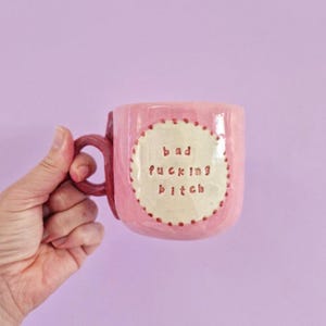 May include: A handmade pink ceramic mug with a glossy finish and a rounded handle. The centre features a cream-coloured circular patch with a scalloped edge, embossed with the text "bad fucking bitch" in dark red, hand-pressed lettering.