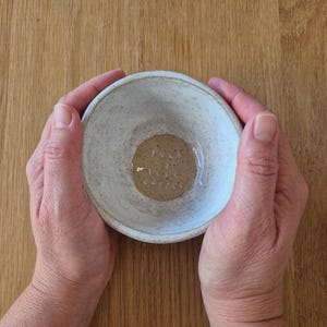 May include: A small, round, off-white ceramic bowl with a speckled texture. The interior base features a circular tan patch with the embossed text "more coffee" in white lettering. The bowl has a thick, uneven rim and a rustic, handmade appearance.