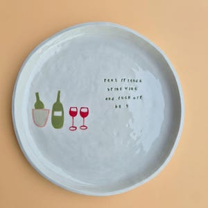 May include: A white ceramic plate with a wide rim, featuring hand-painted illustrations of wine bottles, a wine bucket, and two red wine glasses. The plate has the text "real friends bring wine and f*ck off by 9."