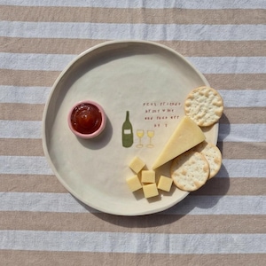 May include: A round, off-white ceramic plate featuring a small pink bowl of red sauce, a wedge of yellow cheese, and several round crackers. The plate is decorated with a green wine bottle, two yellow wine glasses, and the text real friends bring wine and fuck off by j.