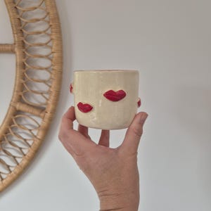 May include: A cream-coloured ceramic mug featuring a smooth, glossy finish and multiple raised, bright red lip motifs applied to the exterior surface. The mug has a cylindrical shape with a flat base and a simple, handleless design.