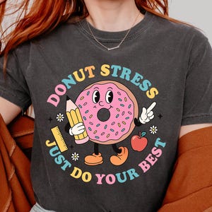 Op de afbeelding: Een grijs T-shirt met een donut-afbeelding en de tekst "Donut Stress Just Do Your Best". De donut is roze met sprankelingen en heeft een glimlachend gezicht. De tekst is in een regenboogkleuren schema.