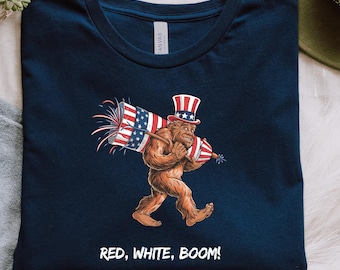 Puede incluir: Una camiseta azul marino con un gráfico de un Bigfoot que lleva un sombrero de tío Sam rojo, blanco y azul y lleva un fuego artificial. El texto "RED, WHITE, BOOM!" está impreso debajo del gráfico.